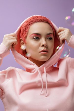 Stylish woman with colored hair in a pink hoodie poses against a violet background surrounded by soap bubbles.